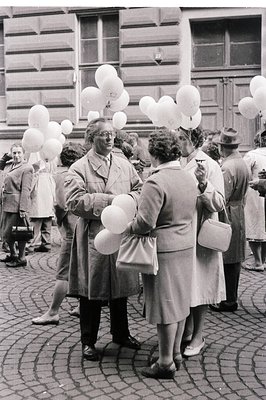 Crowded cobblestone street scene. A man and women stand amid a release of white balloons; onlookers visible. Formal attire su...