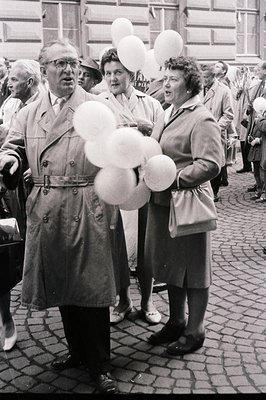 A distinguished gentleman in a trench coat holds a cluster of white balloons, flanked by two women in matching coats, amidst ...