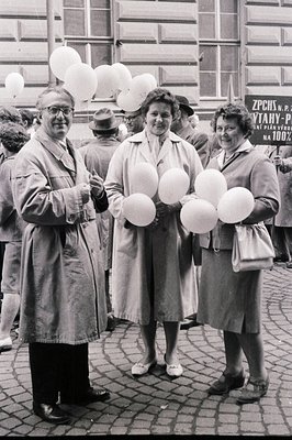 Three formally dressed people holding bunches of pale balloons stand outdoors on a brick-paved street. The group's attire sug...