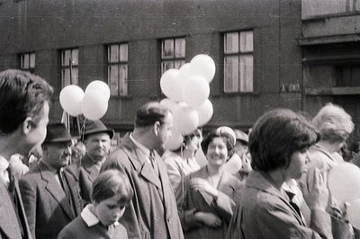 A bustling crowd, primarily men in suits and hats, gather with several bouquets of white balloons. A young boy looks toward t...