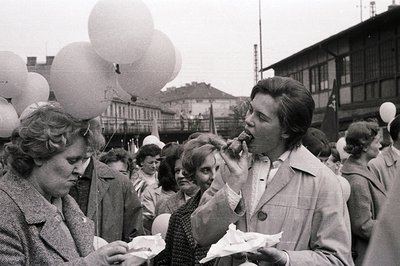 Crowded outdoor scene, likely a public gathering or festival. Individuals hold paper cones filled with food, with one woman p...