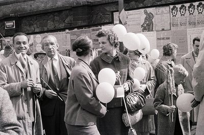 A queue of men and women await something, holding balloons aloft. The scene appears to be in Czechoslovakia, likely Prague, e...