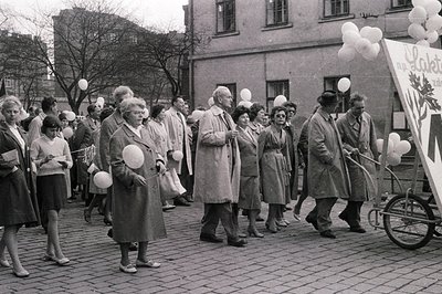 A crowd of formally dressed individuals walks along a street, many holding white balloons. A person pushes a banner on wheels...