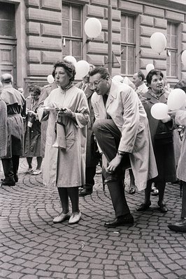 A black and white street scene captures a crowd gathering, with white balloons floating upward. A man in a trench coat and gl...