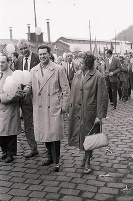 Black and white photograph showing a formally dressed couple walking alongside a crowd of people. Visible are factory chimney...