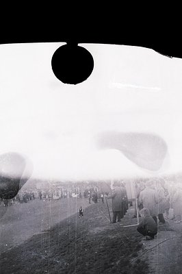 A vintage monochrome photo depicts hot air balloons inflating on a field, attended by a crowd of spectators. Men in period cl...