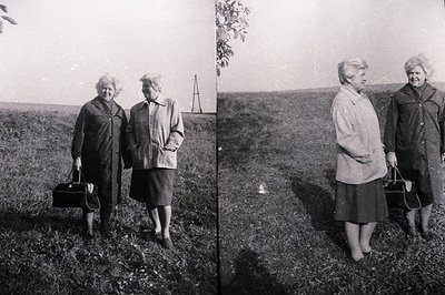 Two women in coats and dresses stand in a field of tall grass. One carries a dark handbag. Possible mid-century portrait, per...