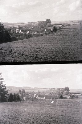 A rural village scene, likely from the mid-20th century. Houses with steeply pitched roofs cluster on a hillside. The image f...