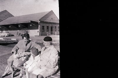 Family portrait: A woman, young child, and military serviceman sit on a bench outside a building resembling a transit station...