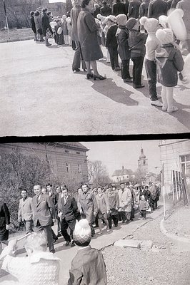 A large group of people, primarily men in suits and hats, is seen walking along a street lined with simple buildings, likely ...