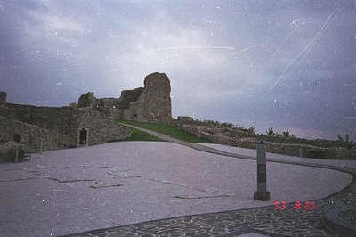 Ruined stone fortifications dominate this view under a cloudy sky. A paved path ascends toward the crumbling walls, bordered ...