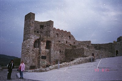 Ruined stone castle wall features crenellations & arched openings. Two figures in modern clothing stand before the structure,...