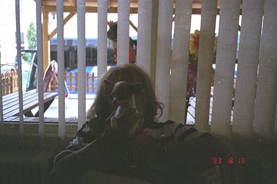 A young person sits indoors, facing away from the camera, partially obscured by vertical blinds. The subject wears a striped ...