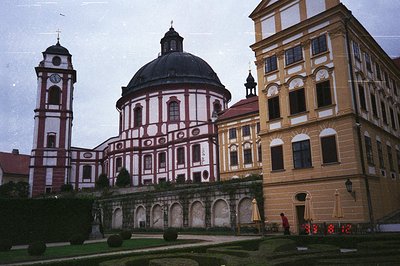 A grand baroque complex featuring a rounded chapel with a central dome, flanked by a tower and a multi-story yellow building....