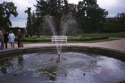 Formal garden scene features a tiered fountain with ducks swimming within a circular pond. A neatly trimmed box hedge and a w...