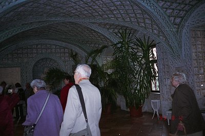 Ornate, arched ceiling of a Moorish-style interior. Three figures in outerwear observe potted palms. Likely a palace or histo...