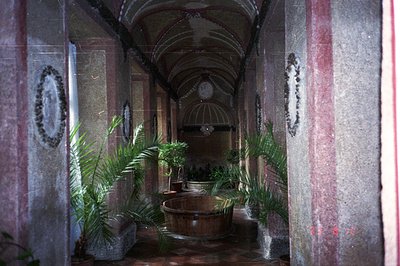 A long, arched hallway leads to a courtyard featuring a large, wooden tub fountain. Decorative moldings and stone pillars def...