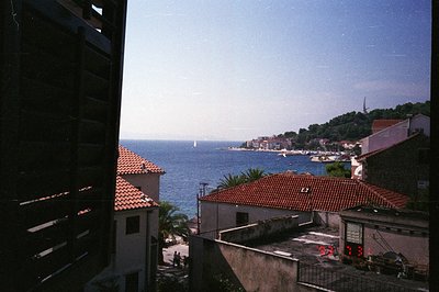 Framed by a dark window mullion, a coastal town view reveals terracotta rooftops, palm trees, and a turquoise sea dotted with...