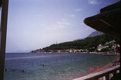 Scenic coastal view of a sandy beach and town nestled against a forested hillside. Swimmers are visible in the turquoise sea....