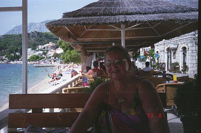 A person sits beneath a thatched-roof veranda overlooking a pebble beach and the Adriatic Sea. Stone architecture and hillsid...