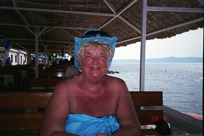 A portrait of a woman seated at a waterfront restaurant, likely on vacation. She wears a blue turban and a matching wrap, rad...