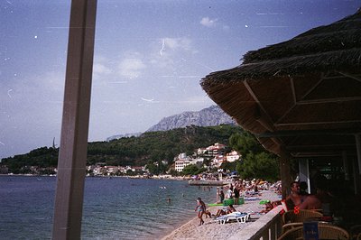 Framed view of a seaside resort, likely the 1970s or 80s. A thatched-roof veranda extends over a sandy beach with sunbathers....