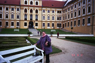 Ornate, yellow stucco facade of a baroque palace features rows of arched windows and a red-tile roof. A tiered formal garden ...
