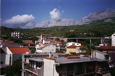 Coastal town view features terraced buildings with red tile roofs, nestled against a backdrop of rugged, forested mountains. ...