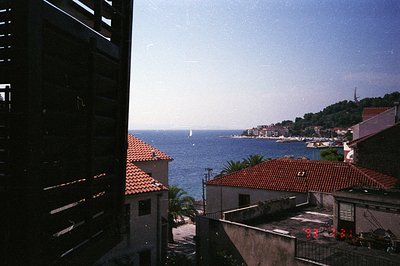 Seascape view through a building's shutters reveals a coastal town with terracotta roofs and a small harbor. Lush green hills...
