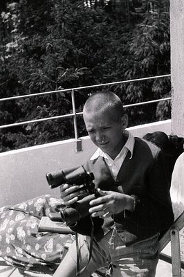 Young boy, likely 1960s-70s, seated and operating a reflex camera, possibly a Nikon or similar model. He wears a short-sleeve...