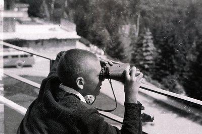 A young boy, likely a child, is depicted looking through binoculars while leaning against a railing. He is wearing a short-sl...