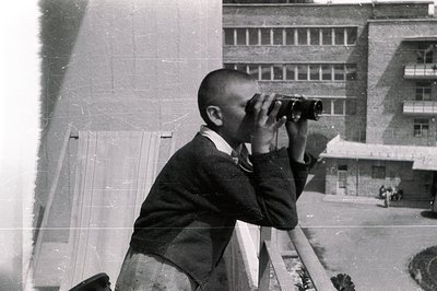 A young boy in a tweed jacket and short pants observes the surroundings through binoculars from a balcony. The building behin...