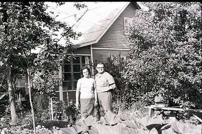 A couple stands before a modest, single-story home with a gabled roof, likely a suburban dwelling. The man wears glasses and ...