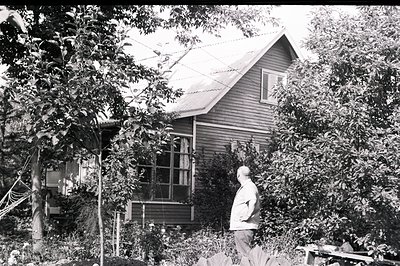 A view of a modestly-sized, clapboard home with a gabled roof, partially obscured by mature trees and foliage. A man, dressed...