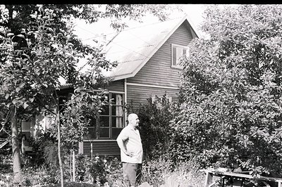A man with a shaved head stands in a lush, overgrown garden before a modest, clapboard house with a steeply pitched roof. The...