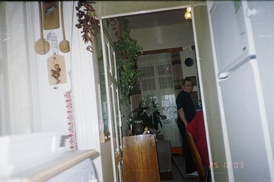 A snapshot captures a domestic interior. Wooden kitchen utensils hang on a wall decorated with floral wallpaper. A man in red...