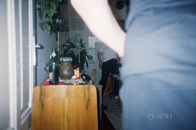 Indoor scene, likely a kitchen or dining area. Features a wooden sideboard with bottled beverages & a potted plant. A blurred...