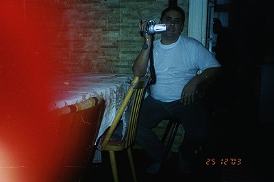 Man in a white t-shirt seated in a wooden chair, operating a video camera. Tablecloth-covered table and exposed brick wall ar...