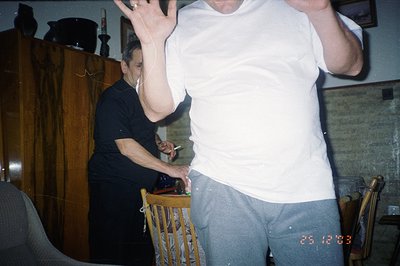 A man in a white t-shirt and grey sweatpants stands with his arms raised, facing the camera. A woman in black is seated at a ...