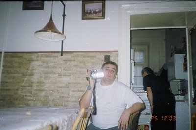 A man sits in a chair holding a vintage film camera. The kitchen features a distinctive tiled backsplash & outdated appliance...