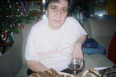 Close-up portrait of a woman seated on a floral-patterned sofa, a Christmas tree visible behind. She wears a white “Eskimo” b...
