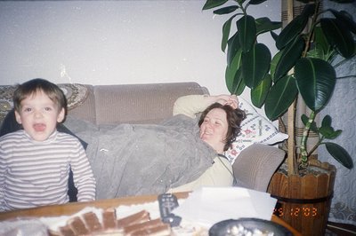 A young boy with a playful expression sits near a woman resting on a textured, beige sofa. The interior features a large ficu...