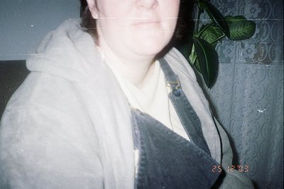 Close-up, likely candid, portrait of a person wearing a hooded sweatshirt over a collared shirt and overalls. A potted plant ...