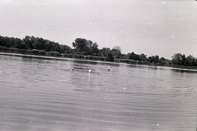 A panoramic view of a lake, possibly a still body of water, with lush vegetation along the banks. Several figures appear to b...