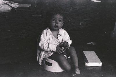 A young child, likely toddler-aged, sits barefoot on a circular object, appearing startled. The subject is centered against a...