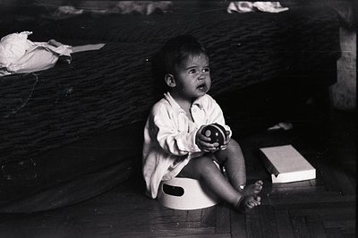 A distressed infant, seated in a plastic potty chair, displays a visible expression of discomfort or frustration. The child w...
