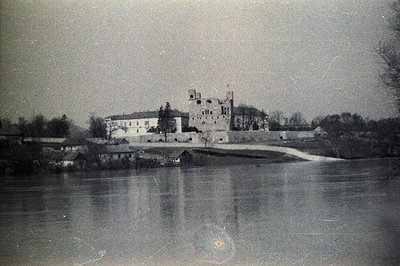 A historic stone castle dominates the landscape, reflected in still water. A narrow bridge leads towards the fortified struct...