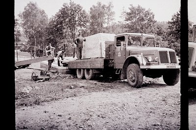 A heavy-duty flatbed truck carries a large, rectangular load—likely construction materials—across a dirt road. Three men are ...