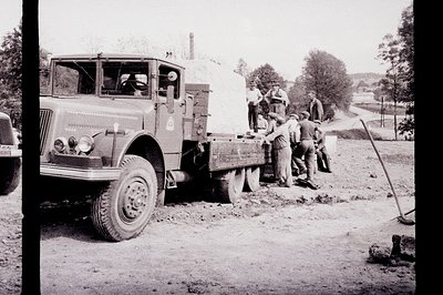 A vintage, heavy-duty truck is pictured, loaded with a large quantity of lumber. Several men, wearing caps, assist in offload...