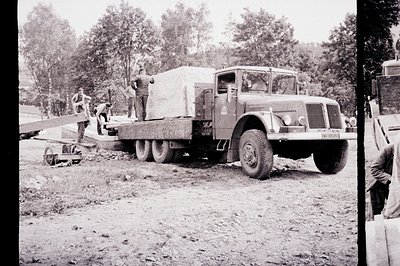 A sturdy, flatbed truck carries a large crate, likely industrial equipment, with several workers visible on the bed and aroun...